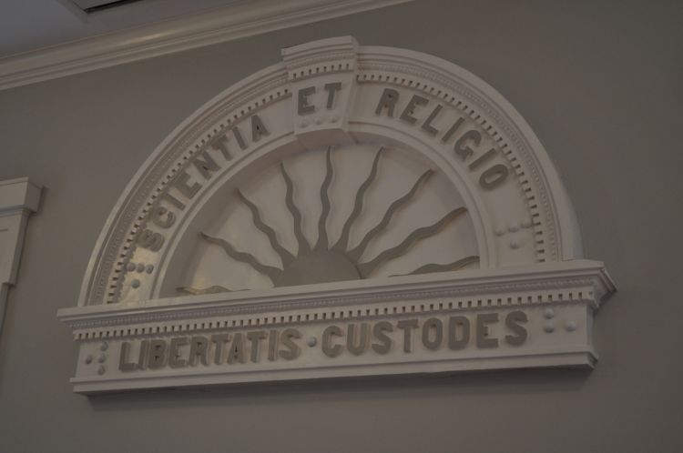 On the west wall hangs a pressed-metal ornamental arch bearing the society’s motto, “Scientia et religio libertatis custodes.”